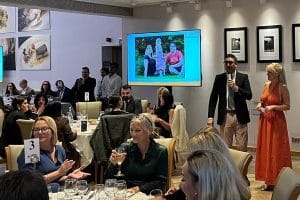 Woman in orange dress and man in suit, stand up to present. People at tables are clapping