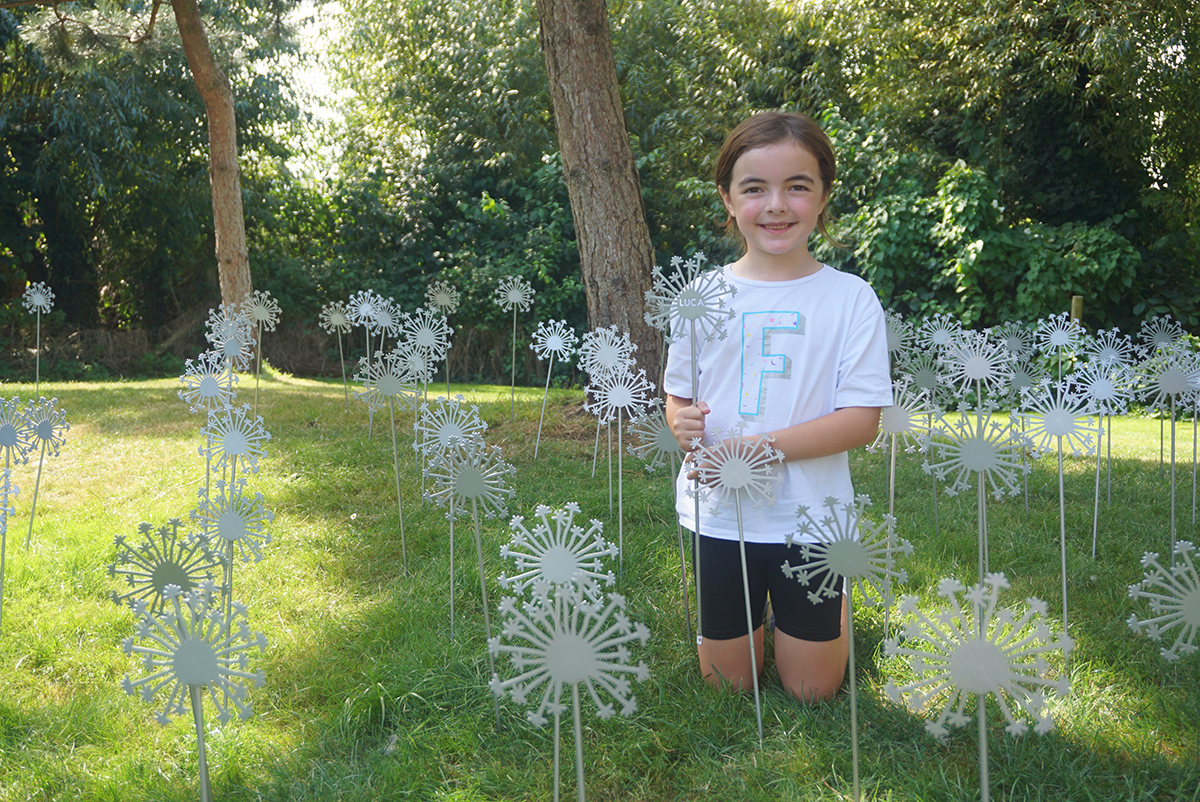 A young girl named Fran holding a sculpted dandelion engraved with her brother's name, Luca, while sitting amongst other sculpted dandelions in a garden.
