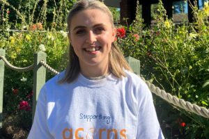 A smiling woman in the Acorns hospice garden, wearing a 'supporting Acorns' t-shirt