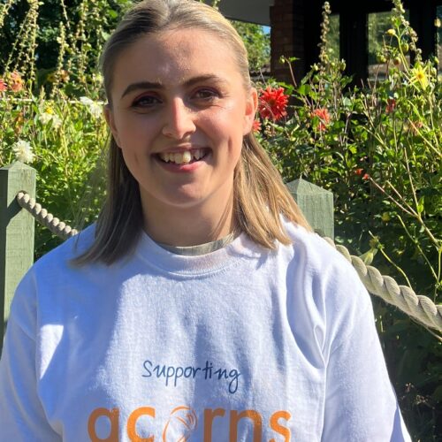 A smiling woman in the Acorns hospice garden, wearing a 'supporting Acorns' t-shirt