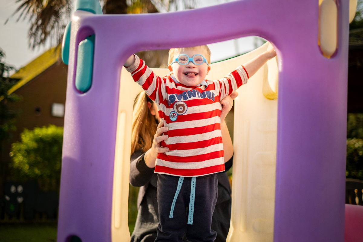 A little boy wearing glasses stands at the top of a small climbing frame, grinning widely
