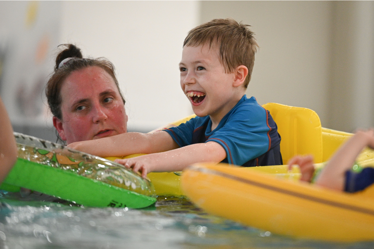 A young boy and a woman in a pool. The young boy is smiling and is in an inflatable seat.