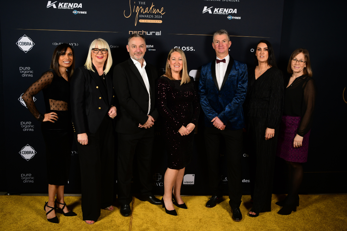 seven people stood in front of a black wall, all dressed in smart black suits and dresses.