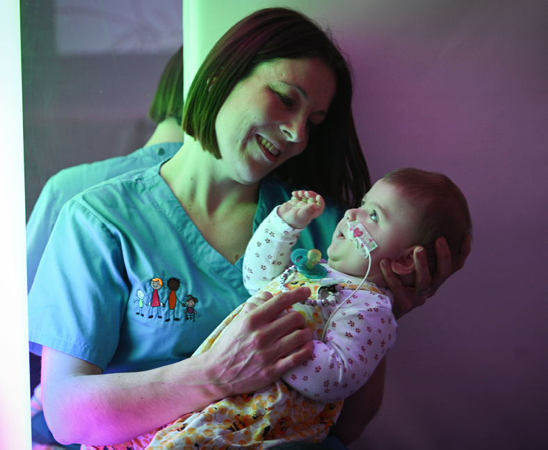 An Acorns Nurse holds a young child in her arms. The child is looking at sensory lights in an Acorns hospice