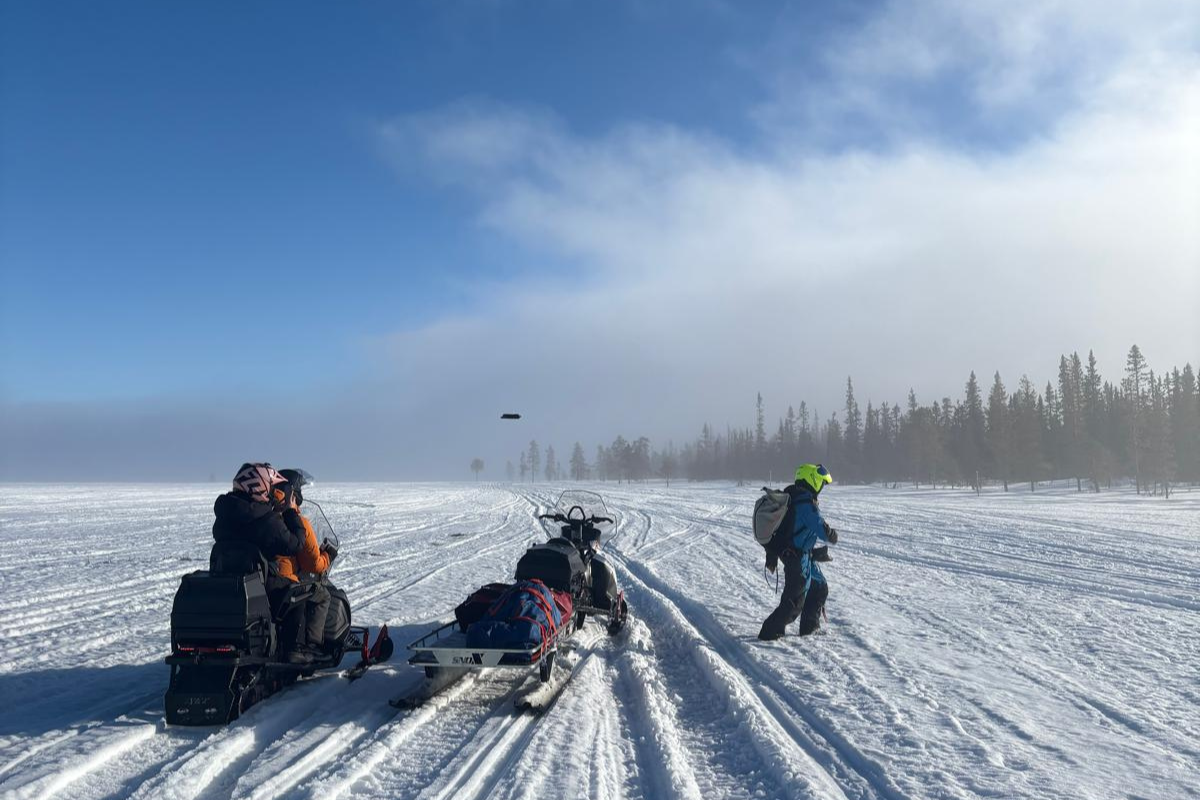 A small group of people, one is walking and two are on snowmobiles on a snowy landscape.