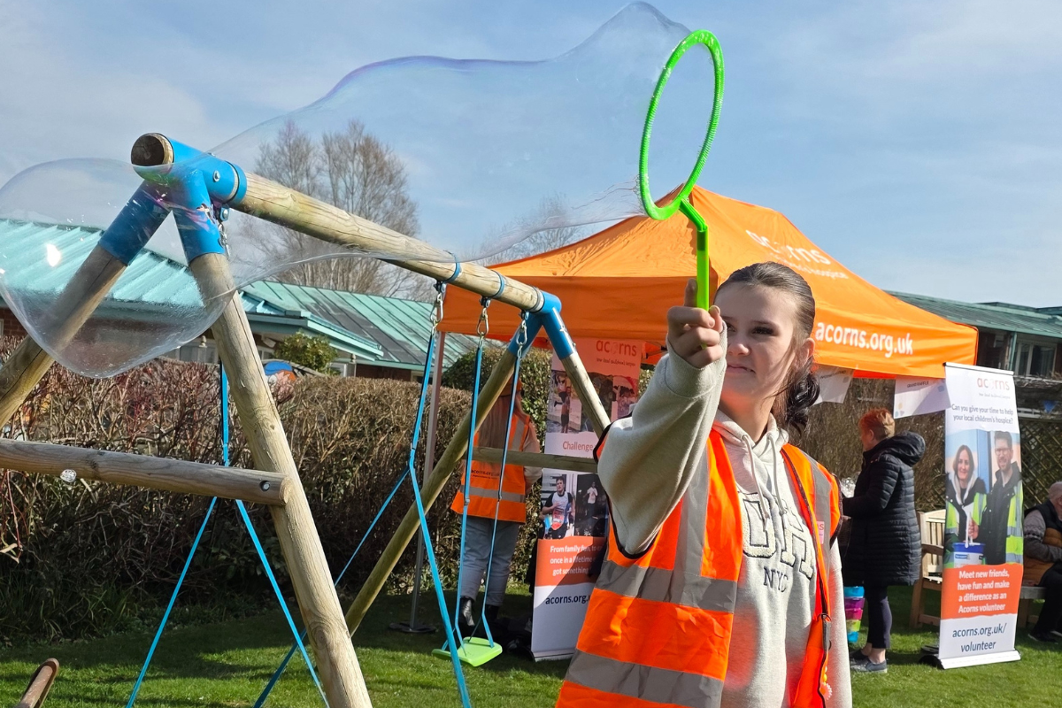 A woman wearing an orange hi vis jacket waving a large stick to create a long bubble.
