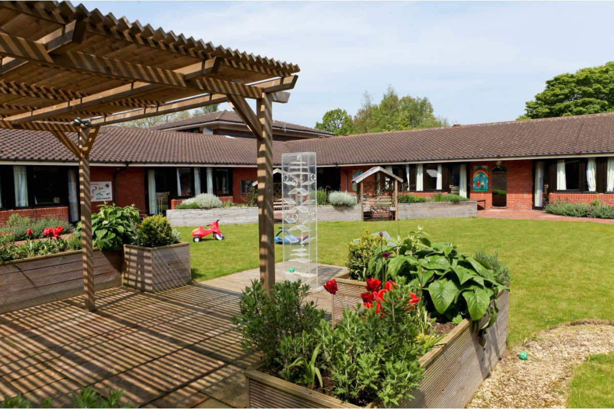 The garden of Acorns Birmingham Hospice which has a small wooden canopy to the left and a large patch of grass to the right and back.