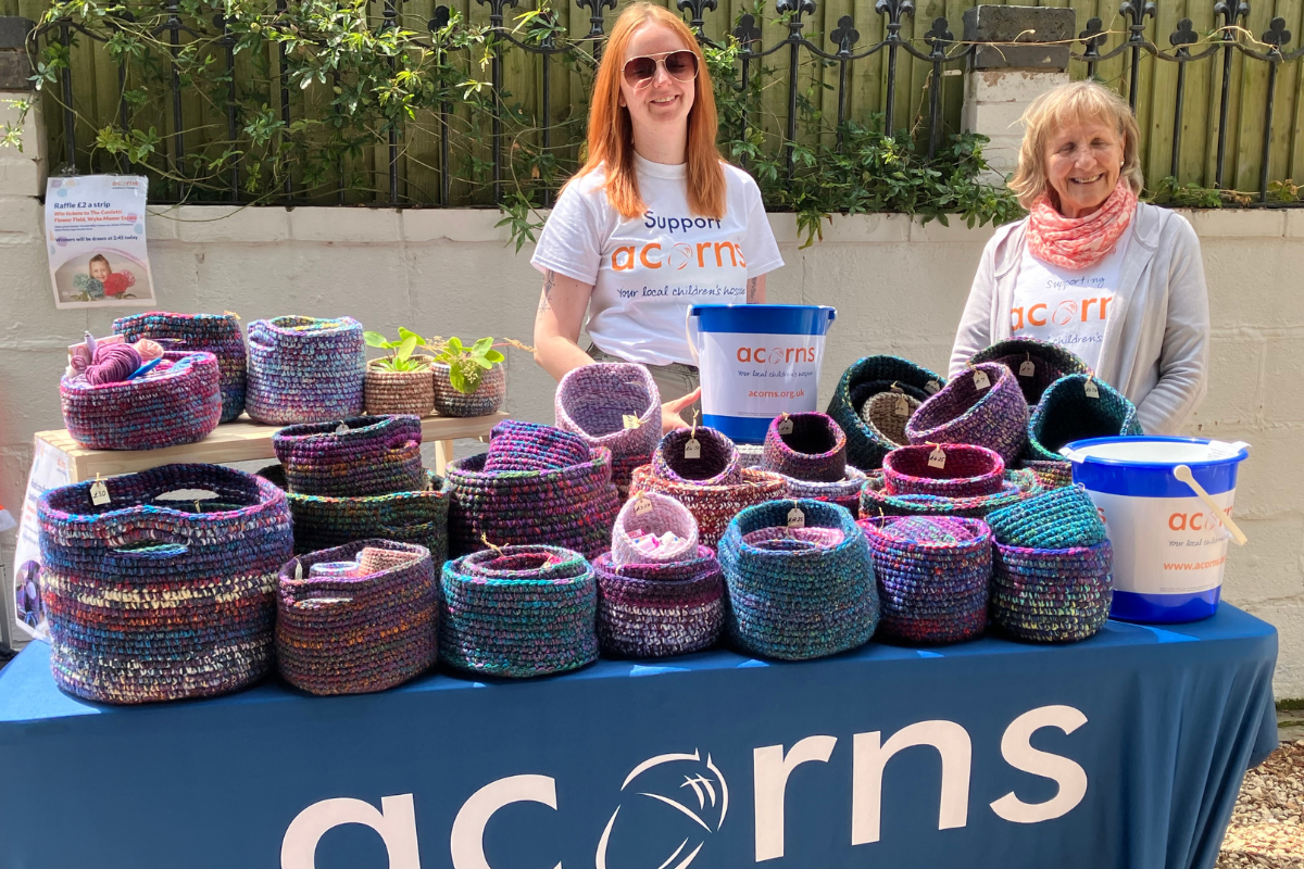 Two women wearing Acorns t-shirt stood behind a stand which has lots of crochet baskets.