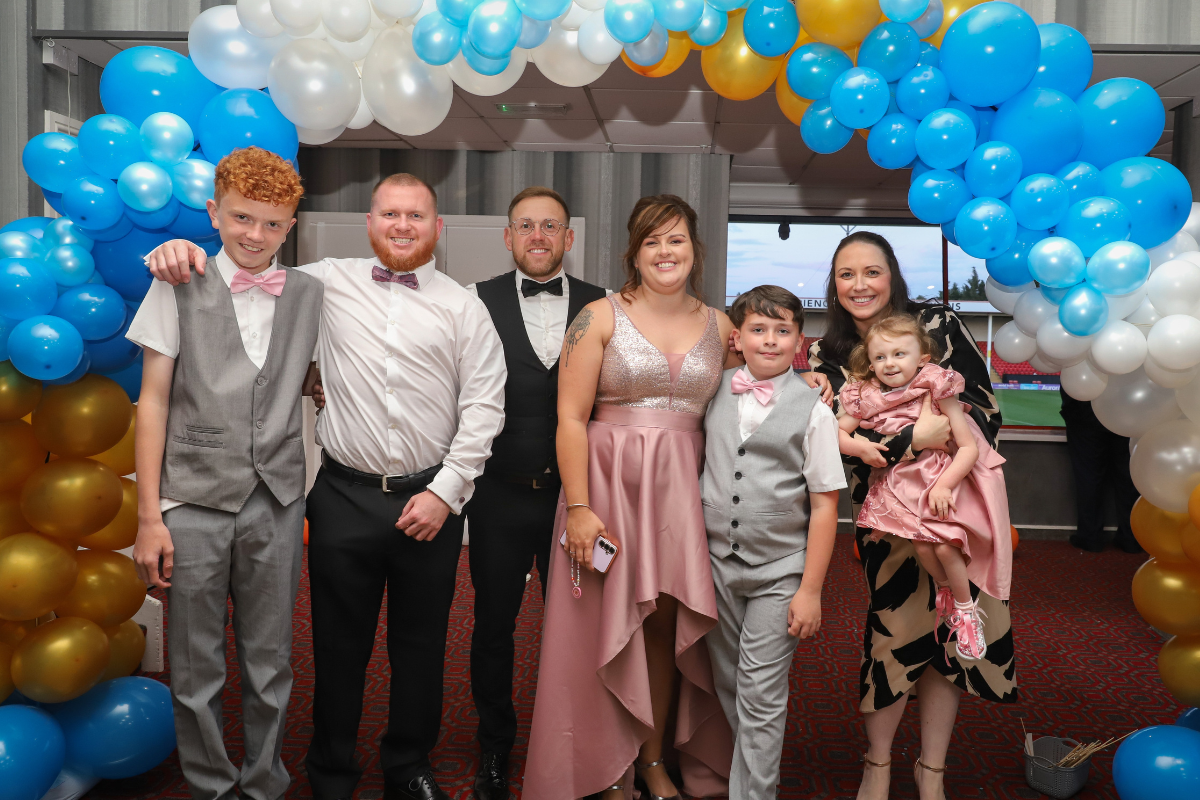 A group of two men, two women and three children all dressed in suits and dresses stood together in front of a balloon arch.