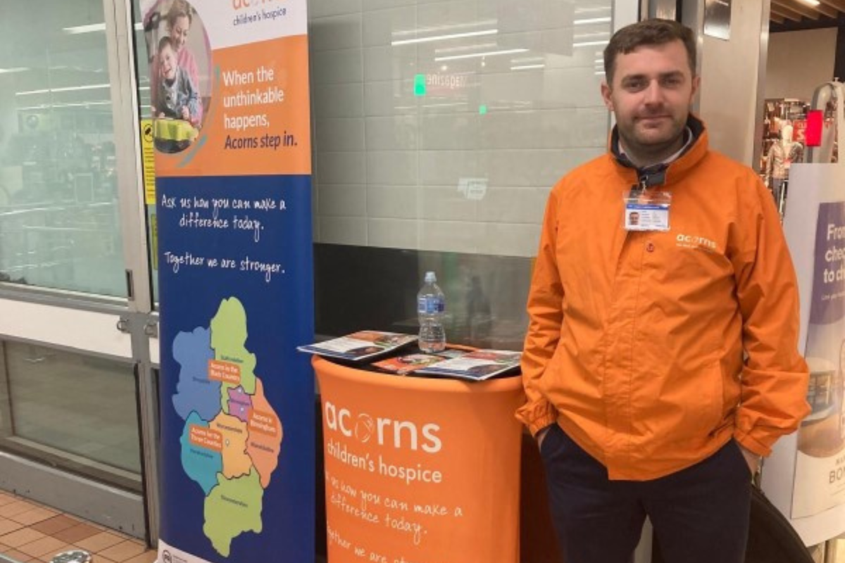 A man wearing an orange Acorns jacket stood next to an Acorns branded banner and stand.