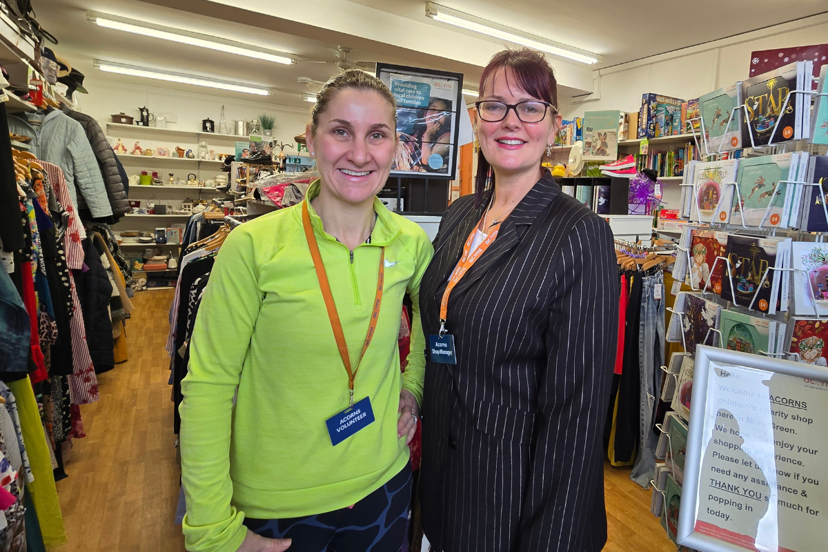 Two woemn stood inside a shop. Hone has glasses and wearing a black blazer and one woman is wearing a green fleece.