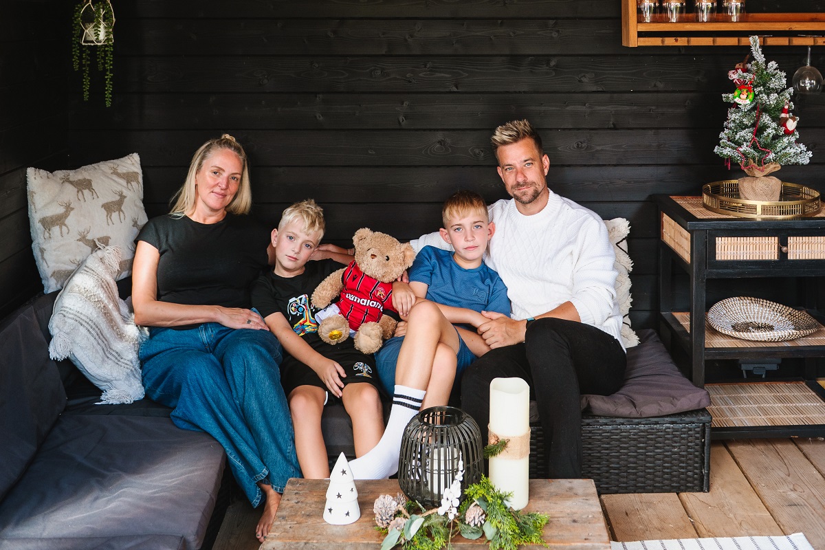 A family with mum, dad and two boys sit on sofa with a teddy bear which is wearing a football shirt. They are surrounded by Christmassy decorations