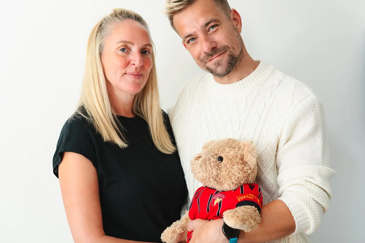 A man and woman stand holding a teddy bear which is wearing a football shirt