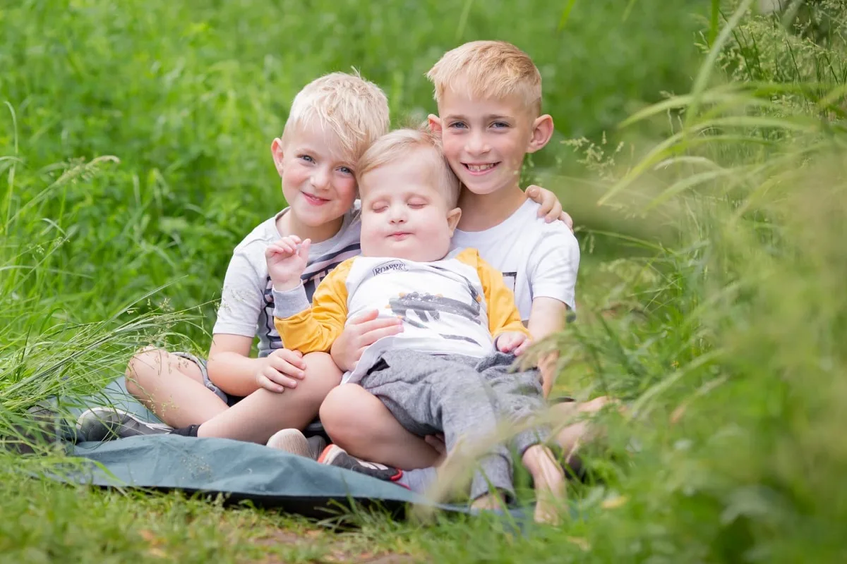 Three boys sit together in a field. The two older are holding the younger one.