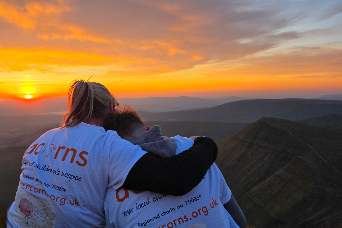 Two people facing away from the camera watching the sunrise over hill, with their arms round each other.