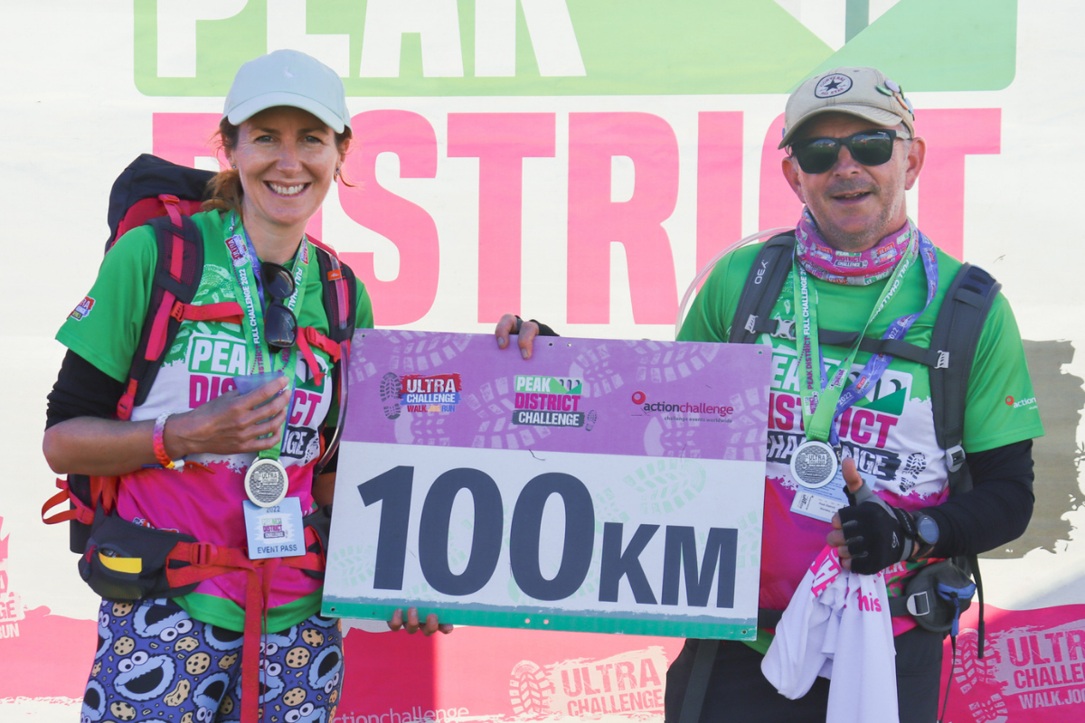 A woman and a man wearing backpacks and medals, they are holding a 100km sign between them.