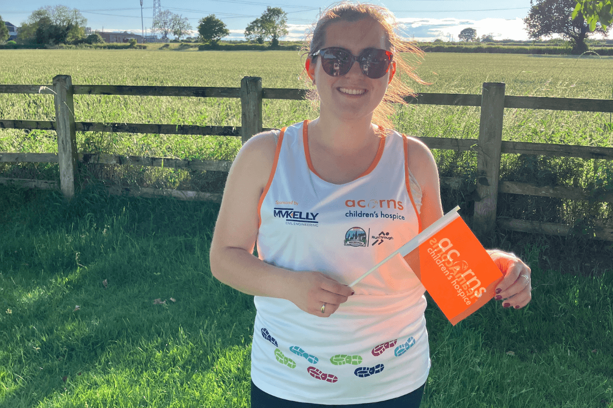 Run Solihull A woman wearing sunglasses and an Acorns Run Solihull running vest. She is standing outside with a field behind her.
