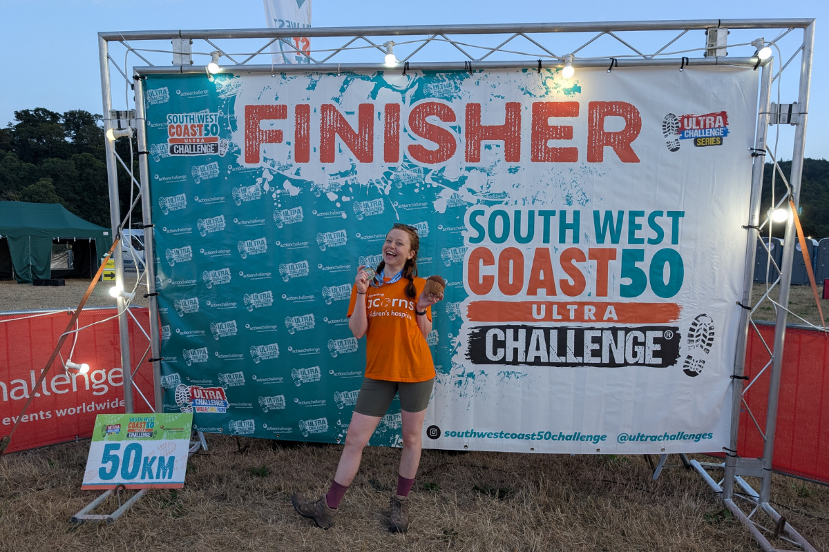 A girl wearing an orange Acorns top with a medal around her neck. She is stood in front of a large South West Coast 50 Challenge finisher banner,