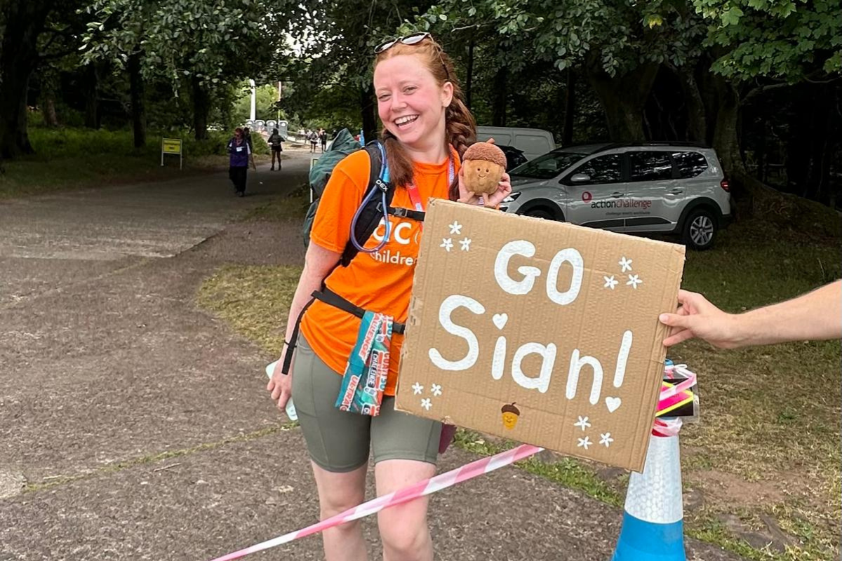 A girl smiling holding a small Acorn teddy in front of a sign that says 'Go Sian' on it.