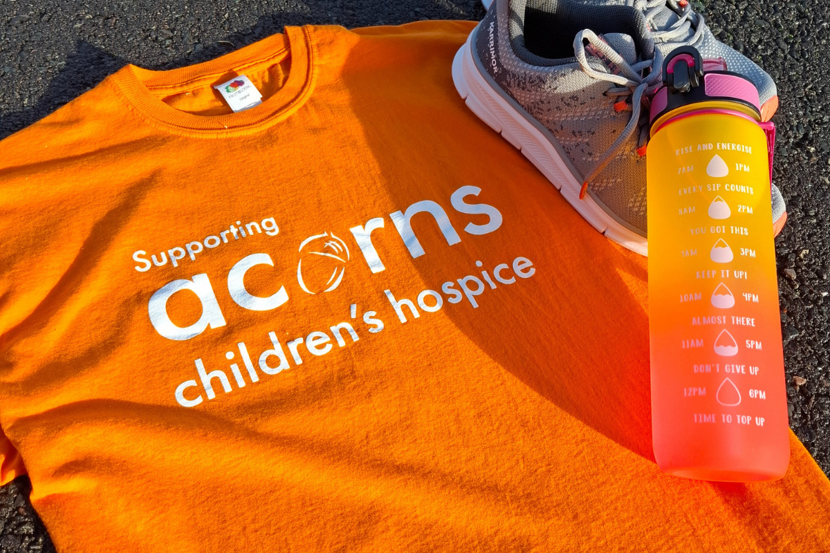 An orange Acorns t shirt laid on the floor with a water bottle and running shoes.