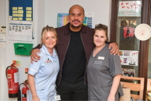 Gabby Agbonlahor at an Acorns hospice smiling with his arm around two Acorns nurses.