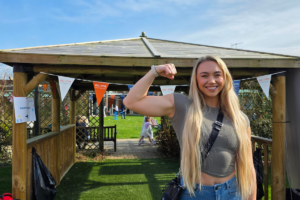 A women with long blonde hair flexing her bicep in front of a garden gazebo.