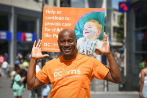 A man wearing an orange Acorns top and holding a large poster about helping to support children's hospice care.