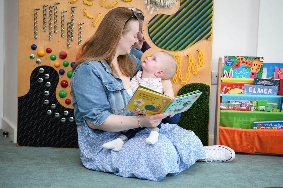 A woman holds a small child in her arms. She's holding a book but they are looking at each other