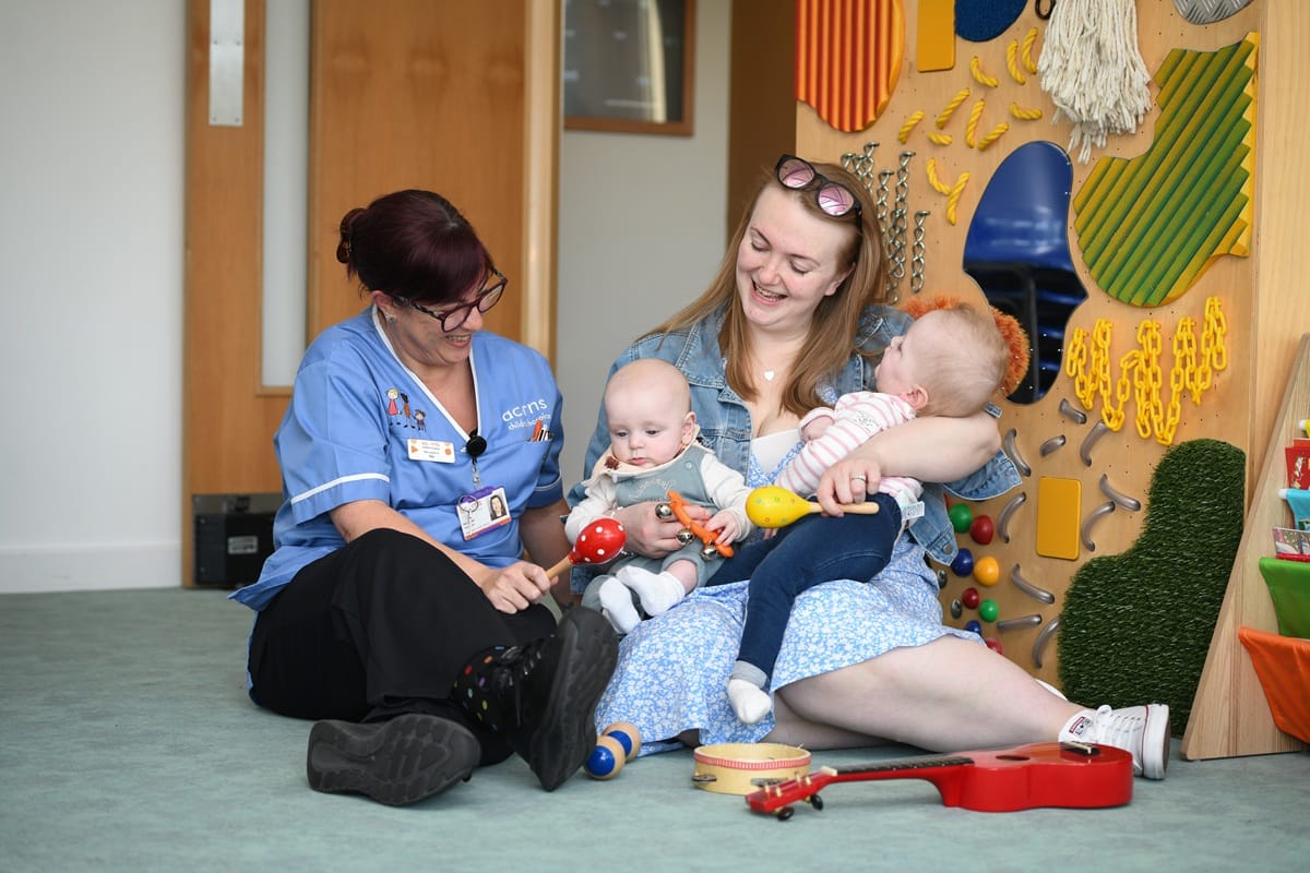 A smiling woman sits next to an Acorns Nurse at an Acorns hospice. She is holding a baby and a young child in her arms and smiling