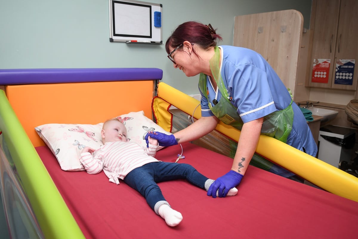 A young child lies on a bed at an Acorns hospice. She is being cared for by an Acorns Nurse