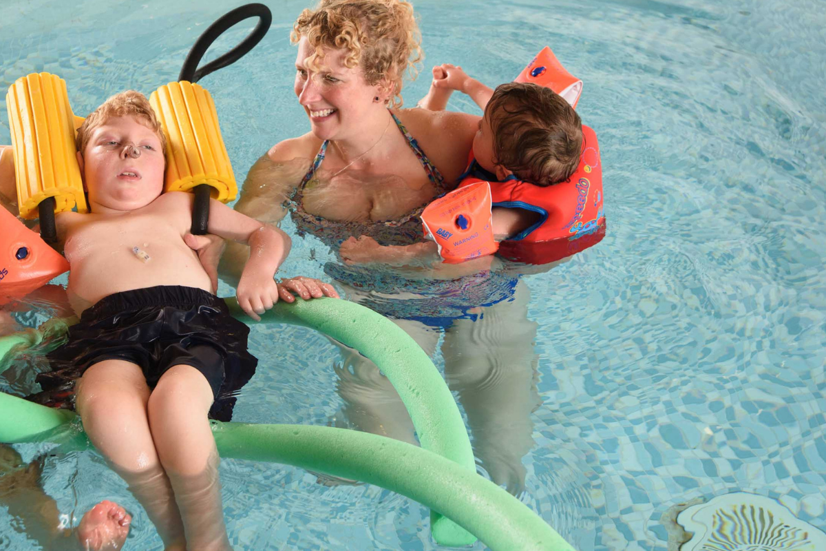 Jack A child in a hydrotherapy pool with lots of floats and a woman next to him holding a small boy in her arms.