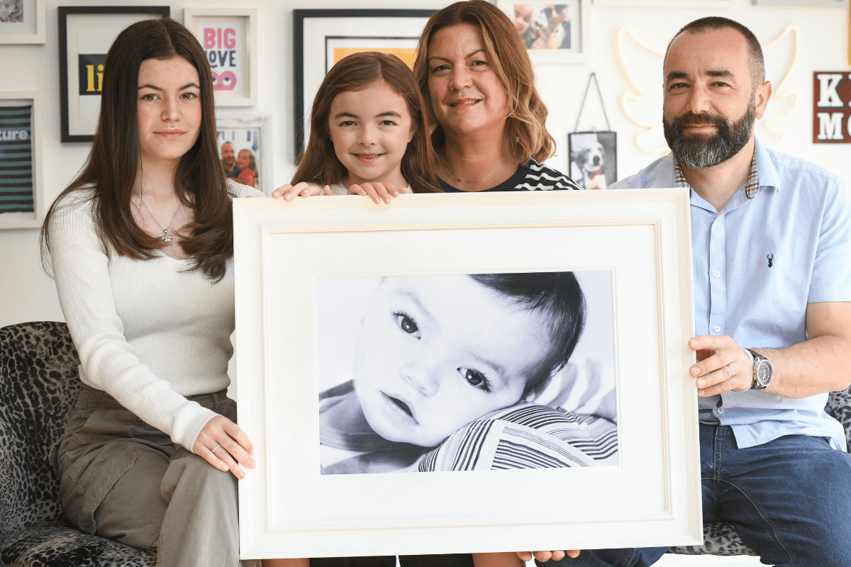 A family with two daughters and their parents sat ona sofa all holding up a large framed photogragh of their late son.
