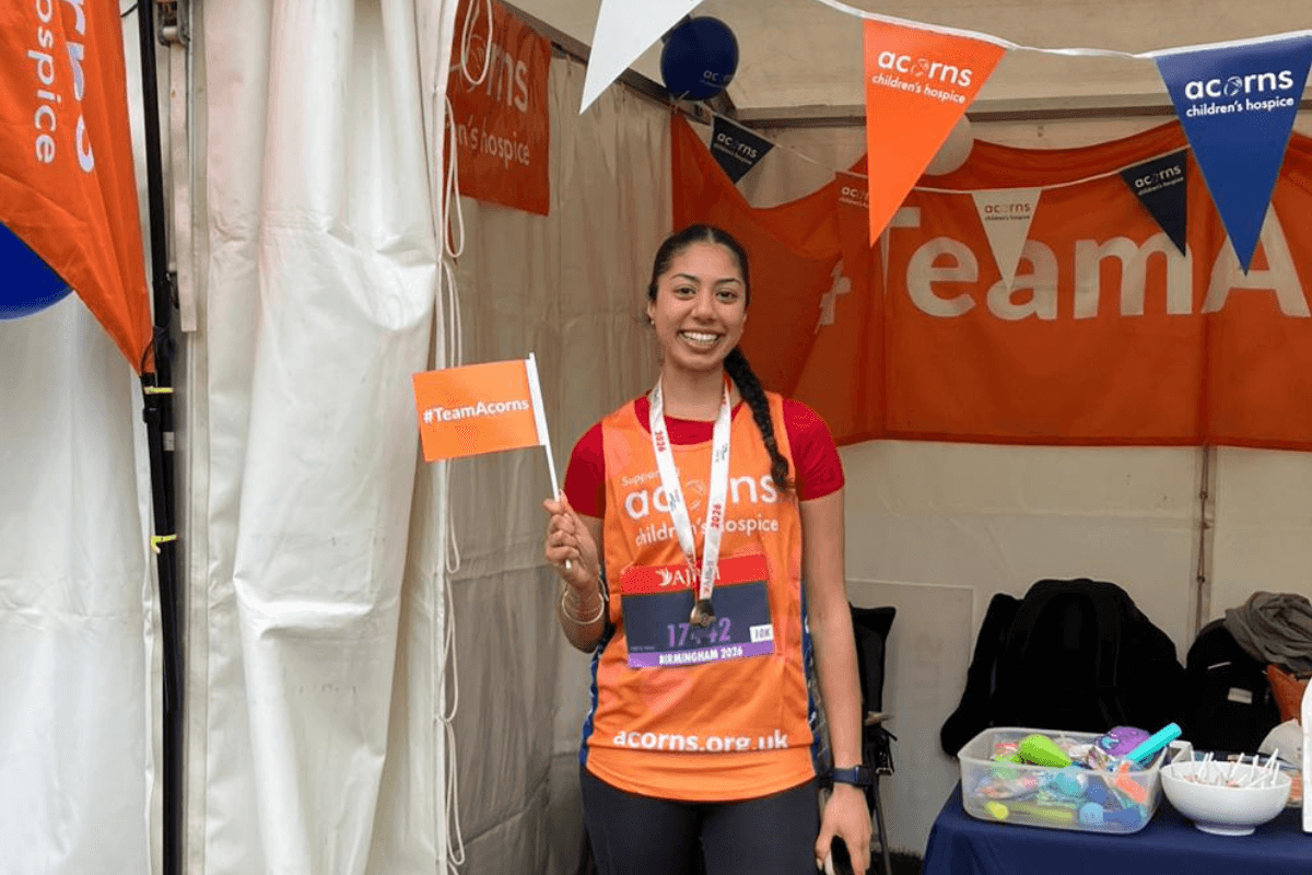 Fundraising A young woman stood underneath a gazebo that is decorate with Acorns bunting and posters. She is wearing an Acorns running vest, a medal and holding a #TeamAcorns flag.