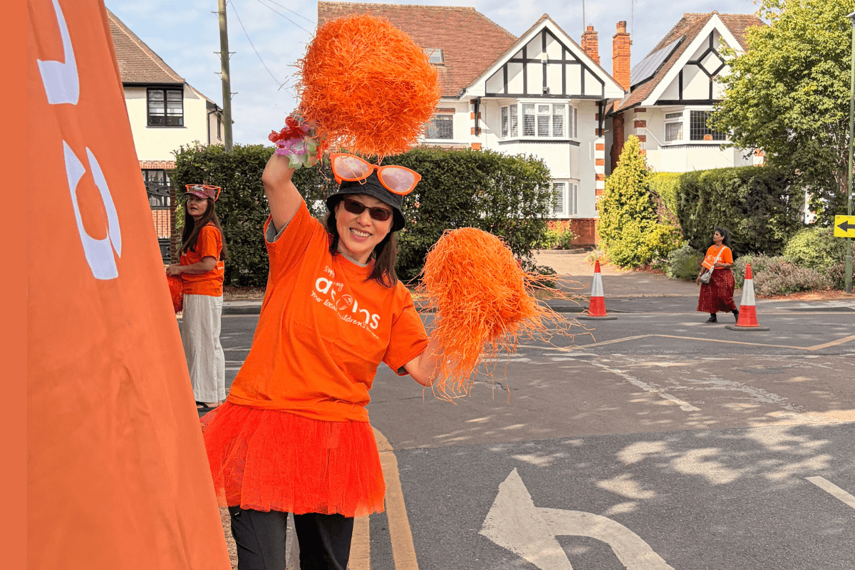 A girl wearing sunglasses and an orange Acorns top, holding up orange pompoms in a cheer.