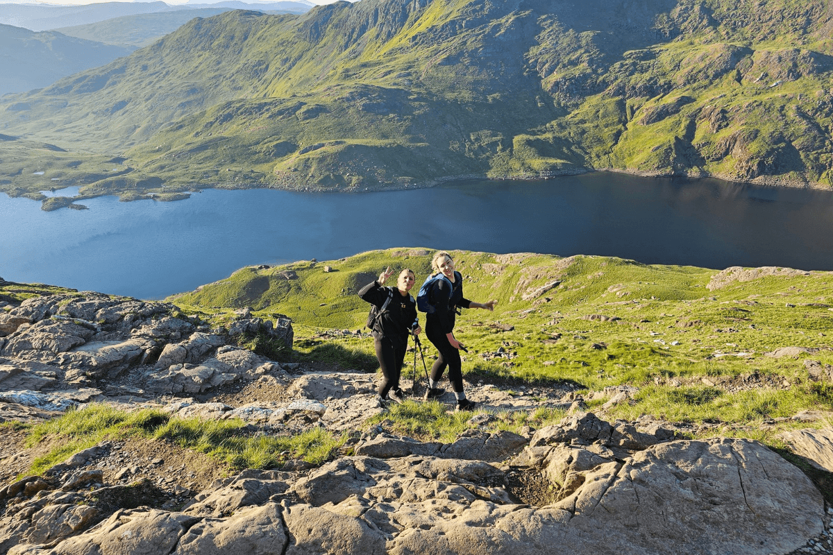 Sunrise trek Two people stood in front of a lake at Snowdon with a grassy hill behind the lake.