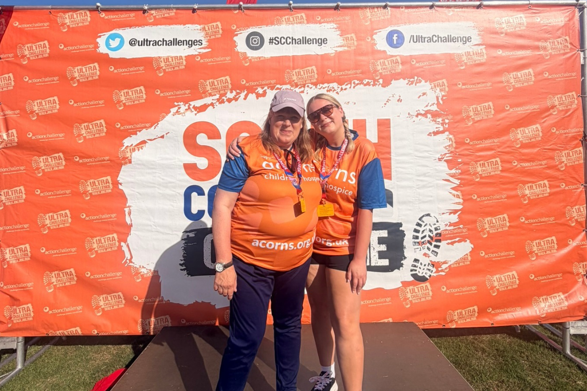 Two woman wearing orange Acorns tops, stood with their arms around each other in front of a large South West Coast banner.